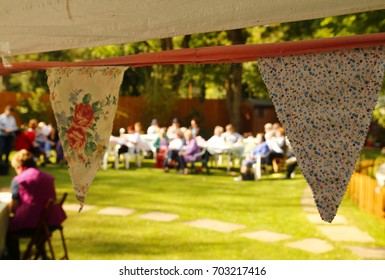 Home-made Fabric Bunting Hangs From A Marquee, With A Soft-focus Background Of Middle Aged And Elderly People Enjoying A Traditional English Garden Party In A Country Village Garden.