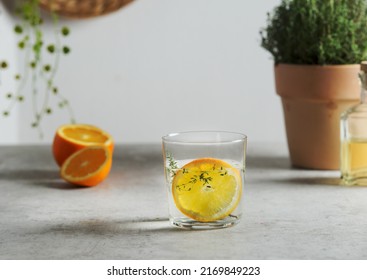 Homemade Flavored Water With Orange Slice And Thyme In Glass At Grey Concrete Table With Fruits And Herbs. Refreshing Summer Drink With Citrus Fruit. Front View.