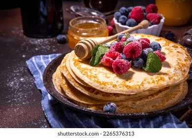 Homemade french tiny pancakes, battercakes with berry. Stack of freshly baked thin crepes on frying pan with fresh blueberry, raspberries and honey. Healthy morning breakfast on dark background