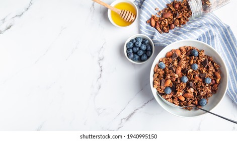 Homemade Granola, Muesli With Pieces Of Dark Chocolate, Nuts And Blueberries In Bowl On White Marble Background. Healthy Breakfast. Top View. Space For Text.