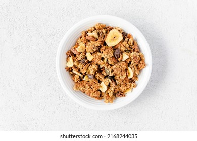 Homemade Granola In A White Bowl On A White Background, Overhead View. Homemade Whole Grain Musli With Bananas And Dark Chocolate For Breakfast. Top View, Light, Airy, Clean. Breakfast Cereal.