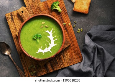 Homemade Green Cream Soup Of Broccoli With Sour Cream And Pumpkin Seeds In A Clay Bowl On A Dark Background. Overhead View, Flat Lay