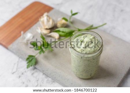 Homemade Green Goddess Dressing in a Jar on a Cement Cutting Board on White Marble Countertop; Parsley and Garlic in Background