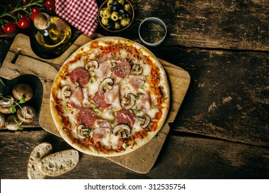 Homemade Ham, Salami And Mushroom Pizza Served On A Board On An Old Rustic Wooden Kitchen Table Surrounded By The Fresh Ingredients From The Recipe, Overhead View With Copyspace
