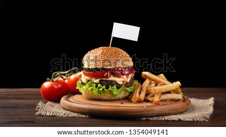 Homemade hamburger close-up with beef, tomato, lettuce, cheese and french fries on cutting board. Small white flag inserted in the burger. Fastfood on dark background.