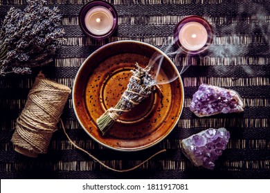 Homemade herbal lavender (lavendula) smudge stick smoldering on brown plate with candles and amethyst crystal clusters for decoration. Spiritual home cleansing concept. Black dark background.