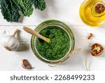 Homemade kale pesto sauce served in a glass bowl over white wooden surface with basic ingredients - kale leaves, walnuts, parmesan, garlic and olive oil