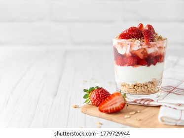 Homemade layered dessert with fresh strawberries, cream cheese or yogurt, granola and strawberry jam in glass on white wood background. Healthy organic breakfast or snack concept. Selective focus.