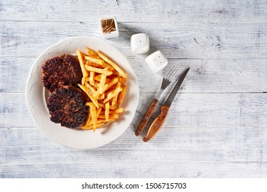 Homemade Meal Of Ground Beef Hamburger Patties And French Fry On A White Plate With Cutlery, Salt, Pepper, And Toothpicks On A White Wooden Background, Top View, Copy Space  