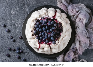 Homemade meringue cake Pavlova with whipped cream, fresh blueberries and blueberry sauce on vintage cake stand on black concrete texture background. Top view, space