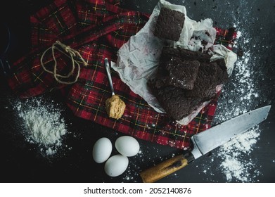 Homemade Moist Dark Chocolate Brownies Cut Into Squares with Peanut Butter Scoop and Christmas Red Checkered Tablecloth and Handmade Knife on Dark Background Top View