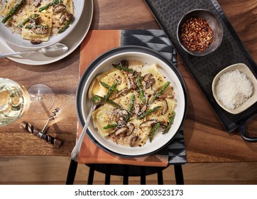 Homemade Mushroom Ravioli With Asparagus In A Bowl On A Wooden Table With A Rust Colored Linen Beneath It. Bowl Has Red Pepper Flakes And Parmesan In Small Bowls Next To It On A Table With White Wine.