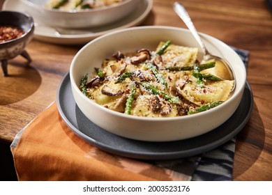 Homemade Mushroom Ravioli With Asparagus In A Bowl On A Wooden Table With A Beautiful Rust Colored Linen Beneath It. Bowl Has Red Pepper Flakes And Parmigiana In Small Bowls Next To It. 