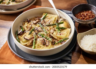 Homemade Mushroom Ravioli With Asparagus In A Bowl On A Wooden Table With A Beautiful Rust Colored Linen Beneath It. Bowl Has Red Pepper Flakes And Parmigiana In Small Bowls Next To It. 