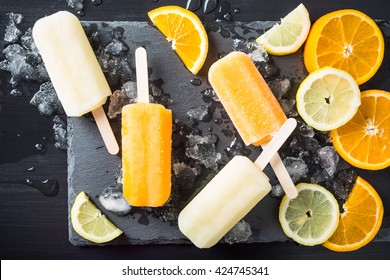 Homemade orange and lemon popsicles with ice and citrus fruits on stone black background. Summer food concept. Top view.