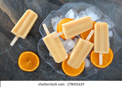 Homemade orange yogurt popsicles in an ice filled bowl with fresh fruit slices against a slate background