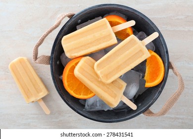 Homemade orange yogurt popsicles in a rustic ice filled tin pail with fresh fruit slices