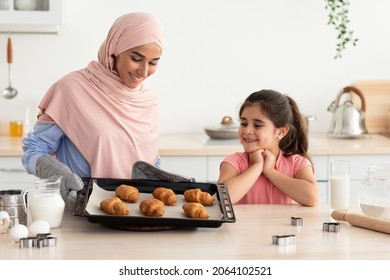 Homemade Pasty. Happy muslim mother in hijab with her little daughter baking together in kitchen, islamic lady holding tray with fresh croissants, cute child looking at tasty snack with excitement