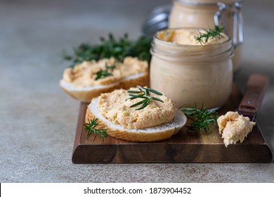 Homemade pate, spread or mousse in glass jar with sliced bread and herbs, light concrete background. Selective focus.
