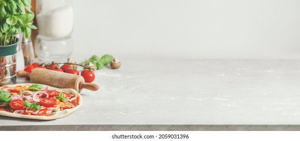 Homemade pizza background with wooden rolling pin, flour and basil leaves on grey kitchen table at white wall background. Cooking Italian food at home. Front view with copy space.  - Powered by Shutterstock