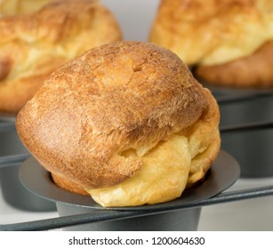 Homemade Popover, Which Is A Puffed, Airy, And Eggy Hollow Roll, Is Fresh From The Oven In A Dedicated Metal Pan With Two More Blurred Popovers In The Grey Background.