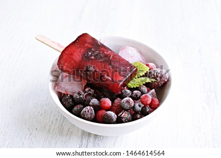 Homemade popsicles of wild berries lie in a bowl full of berries on a bright wooden surface with berries and mint leaves - refreshing healthy organic ice in the hot summer as background