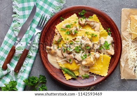 homemade ravioli stuffed with ricotta cheese cooked in creamy garlic mushrooms sauce and served on a plate on a concrete table with napkin and grated parmesan on a paper, close-up