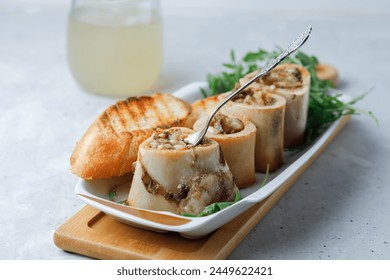 Homemade Roasted Beef Bone Marrow with spoon and croutons on plate, wooden board and beef broth grey background. Side view. Close up.