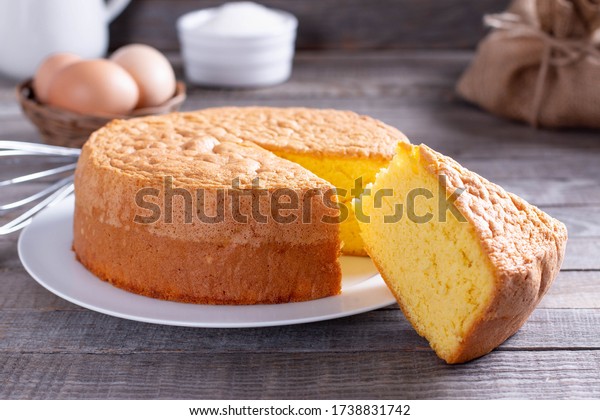 Homemade round sponge cake or chiffon cake on\
white plate so soft and delicious with ingredients: eggs, flour,\
milk on wood table. Homemade bakery concept for background and\
wallpaper.
