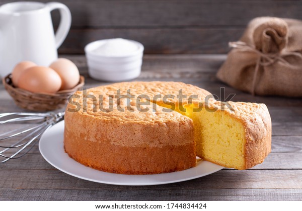 Homemade round sponge cake or chiffon cake on\
white plate so soft and delicious with ingredients: eggs, flour,\
milk on wood table. Homemade bakery concept for background and\
wallpaper.