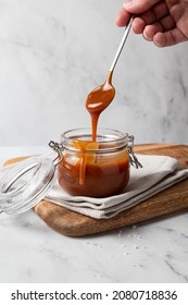 Homemade salted caramel sauce in glass jar on wooden board, grey background. Caramel sauce drips from spoon in hand. - Shutterstock ID 2080718836
