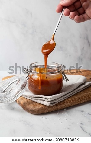 Homemade salted caramel sauce in glass jar on wooden board, grey background. Caramel sauce drips from spoon in hand.