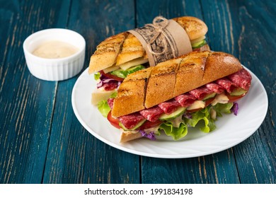 Home-made sandwich of crispy baguette slices with salami, vegetables and lettuce leaves and red cabbage seasoned with a spicy sauce on a white plate and a wooden blue background. 