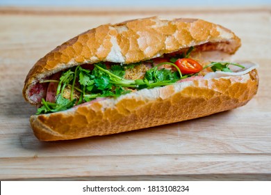 Homemade savoury Vietnamese sandwich - banh mi: Crispy crust of bread baguette filled with coriander, cucumber, chilli, ham, carrot, pickle, pate, green onion, pork displayed on wooden chopped board
