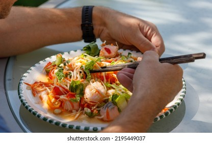 Homemade Seafood Papaya Salad With Prawns And Salmon On A White Plate With Male Hands And Chopsticks, Outdoors In Sun And Shade