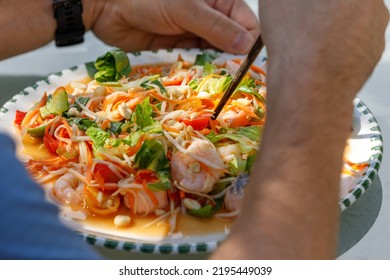 Homemade Seafood Papaya Salad With Prawns And Salmon On A White Plate With Male Hands And Chopsticks, Outdoors In Sun And Shade