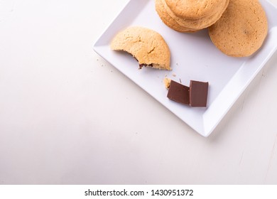 Homemade Stack Of Shortbread Chocolate Cookies With One Bitten Cookie And Two Pieces Of Chocolate On White Plate Wooden Table Top View