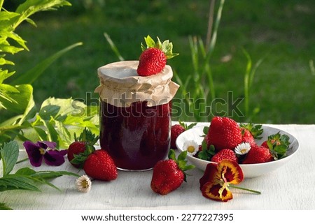 Homemade strawberry jam in glass jar and ripe fresh berries red strawberry with flowers daise and pansy, green leaves on a white wooden table on green natural blur background 