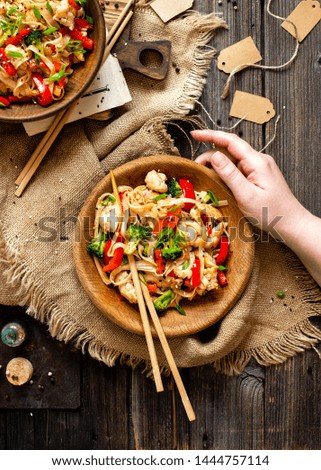 homemade tasty wok noodles with fried chicken fillet, broccoli, red ball pepper, green onion, sesame in two wooden bowls in woman hand on rustic table with sackcloth, chopsticks, spices, top view