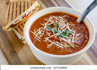 Homemade Tomato And Basil Soup Sprinkled With Parmesan Cheese In White Round Bowl Sitting On Wooden Cutting Board With Grilled Cheese Panini Sandwhich