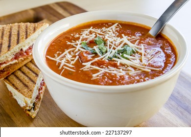 Homemade Tomato And Basil Soup Sprinkled With Parmesan Cheese In White Round Bowl Sitting On Wooden Cutting Board With Grilled Cheese Panini Sandwhich