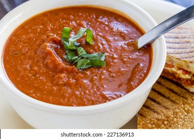 Homemade Tomato And Basil Soup In White Round Bowl With Spoon And Grilled Cheese Panini Sandwich