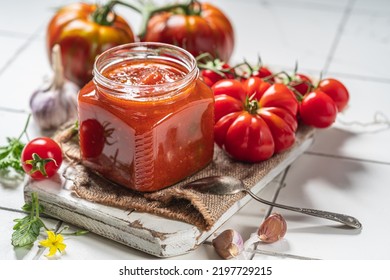Salsa casera de tomate en un frasco de vidrio, tomates y hierbas en su costado. Fondo blanco