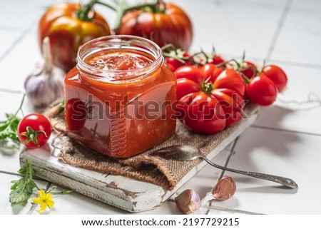 Homemade tomato sauce in a glass jar, tomatoes and herbs on its side. White background