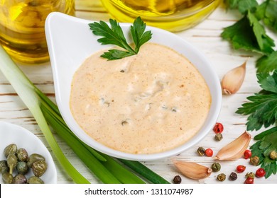 Homemade traditional French remoulade sauce in a White bowl with ingredients-capers, parsley, green onion, mustard, fragrant vinegar, olive oil, garlic on a light wooden table. dip. Selective focus