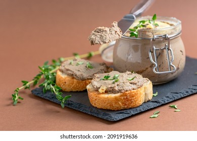 Homemade turkey or chicken liver pate in a glass jar and baguette slices with pate and thyme on a slate serving board, selective focus.