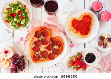 Homemade Valentines Day Dinner. Overhead View Table Scene On A White Wood Background. Heart Shaped Pizza, Pasta, Wine, Cheese Plate And Desserts.