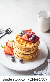 Homemade waffles with berries, powdered sugar and maple syrup on a white plate, gray background.