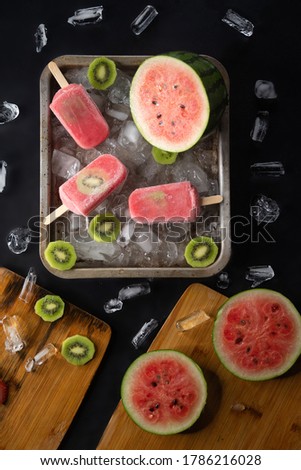 Homemade watermelon and kiwi ice cream or popsicles garnishes with fruits and pieces of ice on black table
