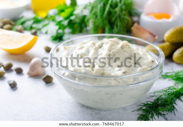 Homemade white sauce tartar tartare with\
ingredients pickles, capers, dill, parsley, garlic, lemon and\
mustard on a light stone background. Horizontal\
image
